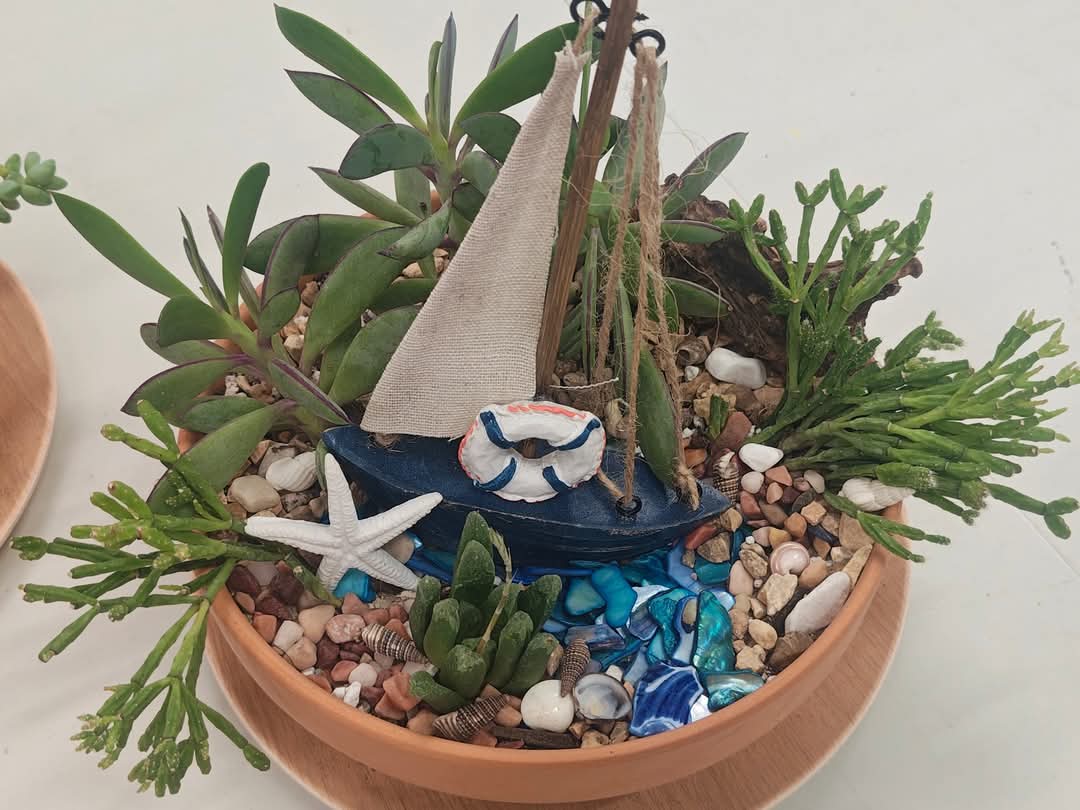 Workshop participant’s succulent garden with a miniature sailboat, shells, stones, and blue glass in a terracotta bowl